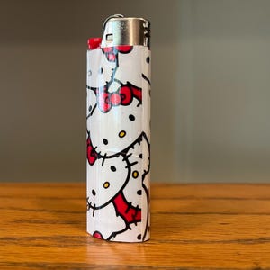 May include: A white lighter decorated with a repeating pattern of a cartoon cat with a red bow. The lighter has a silver metal top and is on a wooden surface.