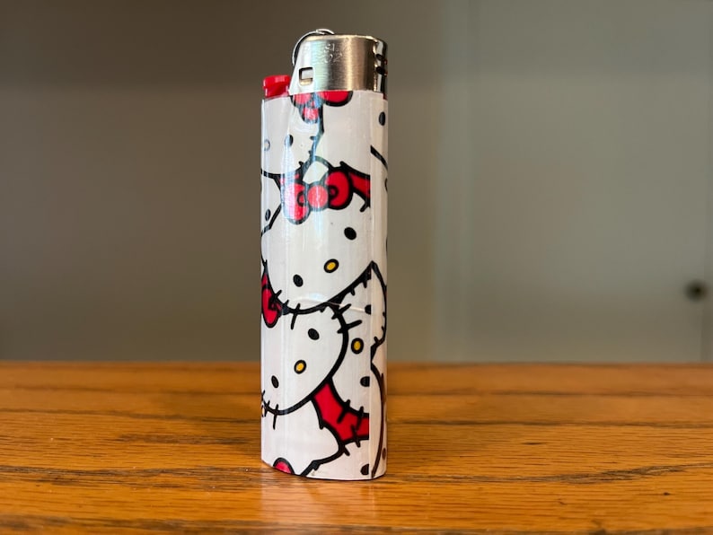 May include: A white lighter decorated with a repeating pattern of a cartoon cat with a red bow. The lighter has a silver metal top and is on a wooden surface.