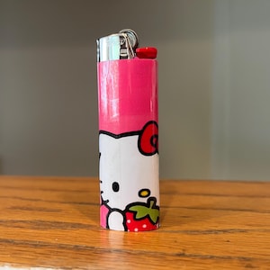 May include: A pink and white lighter with a cartoon character design. The top is silver with a red button. The design includes a red bow, a strawberry, and a smiling face.