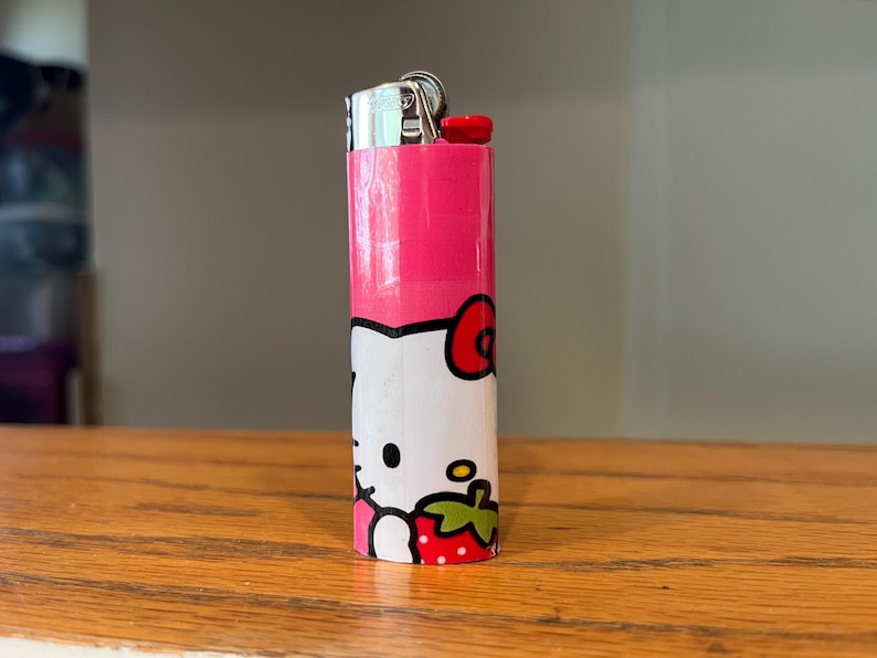 May include: A pink and white lighter with a cartoon character design. The top is silver with a red button. The design includes a red bow, a strawberry, and a smiling face.