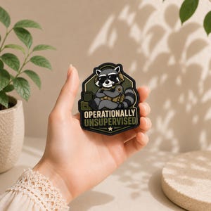 May include: A tactical morale patch featuring a cartoon raccoon wearing sunglasses and a headset, sitting with arms crossed. The patch has a dark green shield shape with the text "OPERATIONALLY UNSUPERVISED" in bold letters and a small star at the bottom.
