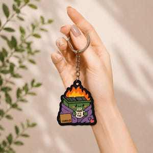 Puede incluir: Un llavero con un contenedor de basura de dibujos animados en llamas, envuelto en una manta morada, sosteniendo una taza con el texto IT'S FINE y un pequeño letrero etiquetado como CHAOS con un corazón. Incluye anilla y cadena metálica.