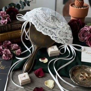 May include: A handheld mirror with a dark bronze finish, adorned with a white lace veil featuring floral embroidery and long white satin ribbons. The mirror is accompanied by two small square ring boxes with gold lettering and a silver dish with gold rings.