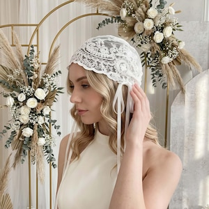 May include: A white lace head covering featuring intricate floral embroidery and scalloped edges. The cap is designed with long, thin white ribbon ties that hang down on the sides, providing a secure fit while adding a delicate detail to the accessory.