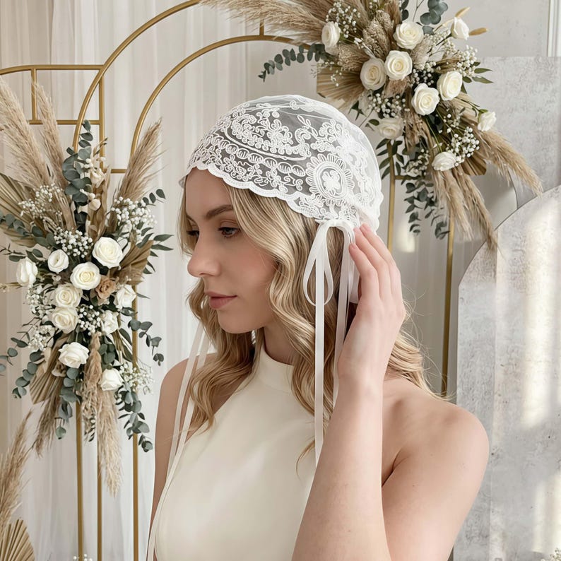 May include: A white lace head covering featuring intricate floral embroidery and scalloped edges. The cap is designed with long, thin white ribbon ties that hang down on the sides, providing a secure fit while adding a delicate detail to the accessory.
