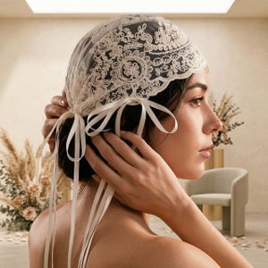 May include: A cream-colored lace head covering featuring intricate floral embroidery and scalloped edges. The accessory is secured with long, matching satin ribbons tied into bows on the sides, with additional ribbon tails hanging down.