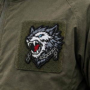 Pode incluir: Uma jaqueta verde-oliva com uma base quadrada de velcro. Fixado na base está um emblema gráfico detalhado de uma cabeça de lobo rosnando com olhos azuis, dentes brancos afiados, uma língua vermelha e uma corrente prateada no pescoço.
