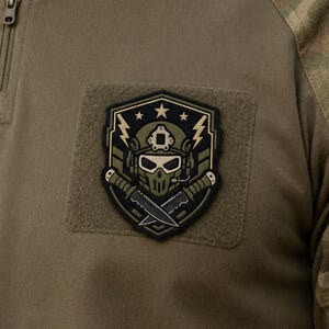 Puede incluir: Una camisa táctica de color verde oliva con un parche en forma de escudo. El parche muestra una calavera con casco táctico y gafas, flanqueada por dos cuchillos cruzados y rayos, todo sobre un fondo oscuro.
