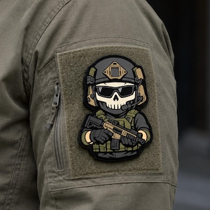 May include: A rectangular morale patch featuring a cartoon soldier with a skull mask, tactical helmet, and sunglasses. The soldier wears olive green tactical gear and holds a rifle. The patch is attached to the sleeve of an olive green jacket.