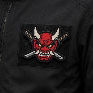 May include: A black fabric jacket featuring a rectangular patch with a red oni mask graphic. The mask has white horns, sharp teeth, and yellow eyes, positioned over two crossed swords with grey blades and yellow and black handles on a black background.