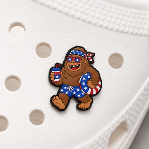 May include: A brown Bigfoot character shoe charm featuring a red, white, and blue patriotic theme. The character wears star-patterned swim trunks, a matching bandana, and sunglasses while holding a drink and a red, white, and blue striped inner tube.