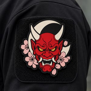 Puede incluir: Un parche cuadrado negro fijado a una prenda negra que presenta una máscara Hannya roja con cuernos y dientes blancos, ojos amarillos y flores de cerezo de color rosa claro dispuestas alrededor de la máscara sobre un fondo de luna creciente blanca.