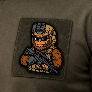 May include: A square morale patch featuring a cartoon illustration of a brown, furry creature wearing a tan tactical helmet with night vision goggles, black sunglasses, and a headset, holding a black rifle, attached to an olive drab fabric.