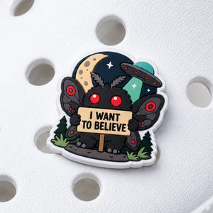 以下が含まれることがあります： I WANT TO BELIEVEと書かれた看板を持つ、赤い目の黒い蛾のような生き物が描かれたシューズチャーム。黄色い三日月、緑色のUFO、暗い緑色の松の木がデザインされており、白いベースの上に配置されています。