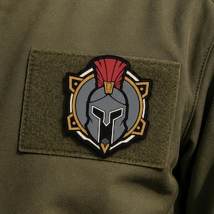 Könnte beinhalten: Ein rechteckiger Aufnäher aus olivgrünem Stoff mit einem zentralen gestickten Emblem eines grauen Spartanerhelms mit rotem Kamm. Der Helm ist von einem kreisförmigen gold-schwarzen Rand mit geometrischen Akzenten umgeben.