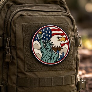 Könnte beinhalten: Ein runder Aufnäher mit der Freiheitsstatue, die eine Fackel hält, einem Weißkopfseeadler und der amerikanischen Flagge vor einer Stadtsilhouette. Das Design verwendet rote, weiße, blaue und beigefarbene Töne mit einem schwarzen Rand.