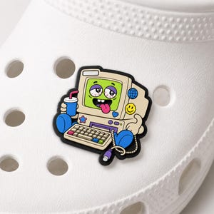 May include: A white shoe charm featuring a cartoon beige computer with a bright green screen, a smiling face with a tongue sticking out, and bloodshot eyes. The computer holds a blue drink and has blue arms and legs with a connected keyboard.