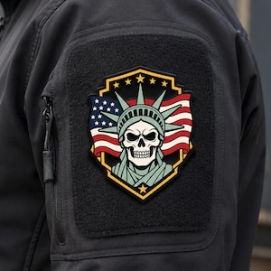 May include: A shield-shaped morale patch featuring a skull wearing a Statue of Liberty crown, set against a stylised American flag background. The design is framed by a gold border with five stars at the top and one star at the bottom, all rendered in a graphic, multi-coloured style.