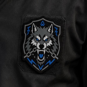 Puede incluir: Un parche de tela negro con un emblema en forma de escudo que muestra una cabeza de lobo gris, ojos azul brillante y la boca abierta mostrando los dientes. El lobo está enmarcado por cadenas grises y rayos azules sobre fondo oscuro.