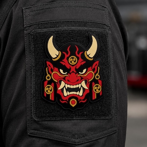 May include: A rectangular black patch featuring a red and black oni mask graphic with cream-colored horns and teeth. The design includes gold circular accents with a triple-swirl symbol, set against a black fabric background with a hook-and-loop backing.