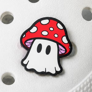 May include: A shoe charm featuring a cartoon-style mushroom with a red cap covered in white spots, a white stem, and a ghost-like face with two black oval eyes. The charm is outlined in black and is attached to a white textured shoe surface.