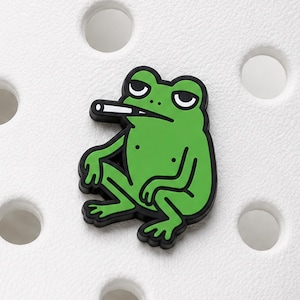 Peut inclure: Un charm de chaussure en forme de grenouille verte avec un contour noir, représentant une grenouille de dessin animé assise avec une cigarette blanche dans la bouche, fixé sur une surface blanche texturée à trous circulaires.