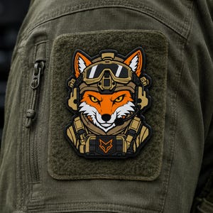 May include: A rectangular morale patch featuring a stylised orange and white fox wearing a tan tactical helmet, goggles, and a headset. The fox is dressed in tactical gear with a small orange fox head logo on the chest plate, all set against a dark background.