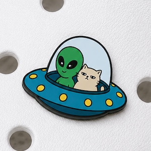 May include: A shoe charm featuring a blue flying saucer with yellow circular accents. Inside the clear dome, a green alien with large black eyes sits next to a beige cat with a grumpy expression. The design is rendered in a flat, cartoon style.