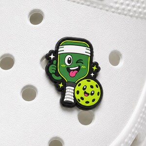 May include: A shoe charm featuring a cartoon pickleball paddle with a green face, white headband, and a winking expression, positioned next to a smiling yellow pickleball with black holes, all outlined in black on a white perforated shoe.