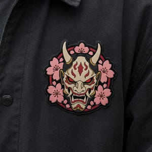 Könnte beinhalten: Ein kreisförmiger Aufnäher mit einer stilisierten japanischen Hannya-Maske mit cremefarbenen Hörnern, roten Akzenten und scharfen Zähnen, umgeben von einem Kranz aus hellrosa Kirschblüten vor einem dunkelroten Hintergrund auf einer schwarzen Jacke.