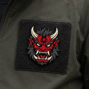 Puede incluir: Un parche táctico cuadrado de color negro con un gráfico detallado en relieve de una máscara de demonio japonesa roja y gris, con colmillos blancos afilados y cuernos curvados. El parche está sujeto a una chaqueta de color verde oliva.
