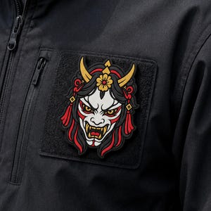 May include: A rectangular black patch featuring a detailed graphic of a Hannya mask with white skin, red accents, yellow horns, and yellow eyes. The mask is adorned with a yellow flower at the top and red tassels on the sides, attached to a black jacket.