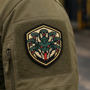 Puede incluir: Un parche con forma de escudo que muestra una cabeza de dragón verde central con ojos rojos y lengua bífida, rodeada por varias cabezas de serpiente más pequeñas. El diseño es negro, verde, rojo y beige sobre una manga de tela verde oliva.