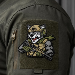 Puede incluir: Un parche táctico que muestra un lobo de dibujos animados con casco de combate color canela, auriculares y bufanda verde. El lobo gruñe y sostiene un rifle negro con silenciador. El parche está sujeto a la manga de una chaqueta verde oliva.