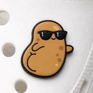 Op de afbeelding: Een beige, aardappelvormige schoenbedel met een stripfiguur die een zwarte zonnebril draagt. Het figuurtje heeft een kleine glimlach, bruine vlekjes en een zwarte omlijning, bevestigd op een witte, gestructureerde schoen met gaten.