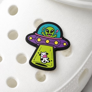 May include: A shoe charm featuring a purple UFO with yellow lights and a teal cockpit containing a green alien. A bright green tractor beam extends downward from the UFO, capturing a small black and white cow. The charm is attached to a white shoe.
