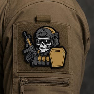 May include: A tactical morale patch featuring a stylised skull character wearing a combat helmet, goggles, and headset. The character holds a rifle in one hand and carries a tan shield in the other, with tan tactical gear pouches visible.