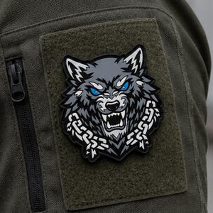 May include: A tactical morale patch featuring a stylized gray wolf head with piercing blue eyes and bared teeth, surrounded by a white chain link design. The patch is attached to a dark olive green fabric surface with a black zippered pocket.