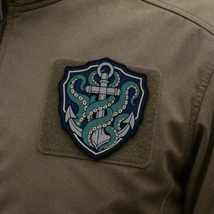 Pode incluir: Um casaco bege com um emblema em forma de escudo no peito. O emblema apresenta uma âncora cinzenta entrelaçada com tentáculos de polvo azul-petróleo sobre um fundo azul escuro, tudo envolvido por uma borda azul-marinho numa base bege.