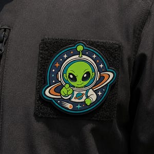May include: A circular rubberized patch featuring a green alien character wearing a white space helmet and suit. The design includes a dark blue background with orange planet rings, white stars, and a small comet. The patch is attached to black fabric.