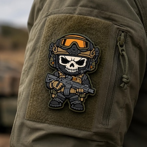 May include: A tactical morale patch featuring a cartoon-style skull character wearing a tan helmet with orange goggles and a black face mask. The character is dressed in dark tactical gear and holds a rifle, rendered in shades of black, tan, and orange.