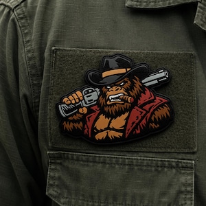 May include: A rectangular morale patch attached to an olive green fabric garment. The patch features a graphic illustration of a brown gorilla wearing a black cowboy hat and a red jacket, holding a shotgun over its shoulder while smoking a cigar.