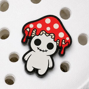 May include: A white enamel pin shaped like a small creature with a red mushroom cap featuring white spots. The mushroom cap has a dripping effect, and the creature has large black circular eyes and a stitched mouth, outlined in black metal.