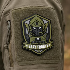 May include: A tactical morale patch featuring a stylised character in a hooded combat helmet and mask, set against a dark shield background. The design includes olive green and black tones with the white text STAY FROSTY on a banner at the base.