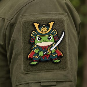 May include: A rectangular morale patch featuring a cartoon frog dressed in green and red samurai armor with a gold helmet crest. The frog holds a katana sword with a white blade. The patch is attached to an olive green fabric sleeve.
