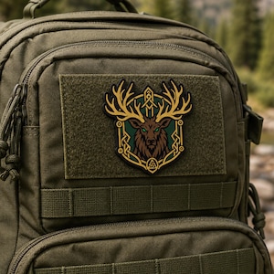 May include: An olive green tactical backpack featuring a rectangular patch with a brown deer head illustration. The deer has large antlers, glowing green eyes, and is framed by a gold-colored border with intricate knotwork and a small triangle symbol.