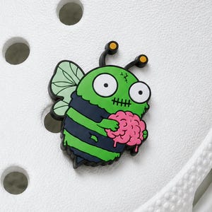 May include: A shoe charm featuring a cartoon zombie bee with bright green skin, black stripes, and light green wings. The character has large white eyes, black antennae with yellow tips, and a stitched mouth, while holding a pink brain in its hands.