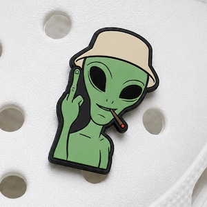 Puede incluir: Un adorno para calzado con un personaje alienígena verde que lleva un sombrero de pescador beige. El alienígena tiene ojos negros grandes, una boca pequeña con un cigarrillo marrón y muestra el dedo medio. El adorno tiene un borde negro.