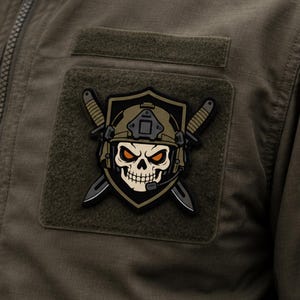 May include: A tactical morale patch featuring a skull wearing a combat helmet with a headset, centred on a shield emblem. Two crossed combat knives are positioned behind the shield. The design uses olive green, black, and orange accents.