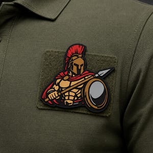 May include: An olive green polo shirt featuring a rectangular hook-and-loop patch on the chest. The patch displays a graphic of a warrior wearing a bronze helmet with a red crest, a red cape, and holding a spear and a round shield in black and gold.